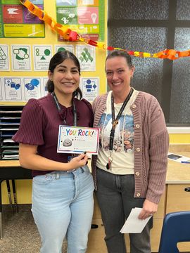 Two women smile in a classroom, holding a certificate in front of colorful wall displays.