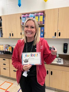 Woman in pink jacket holding “You Rock!” sign in a classroom, smiling beside cabinets and counters.