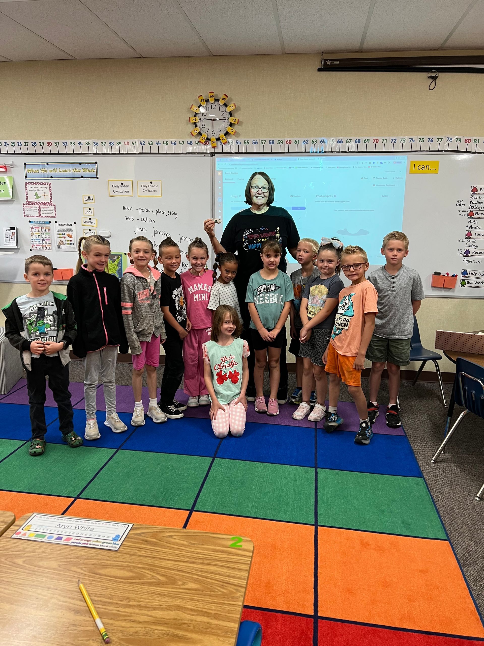 Classroom group photo of children standing with a teacher on colorful floor mats in front of a whiteboard
