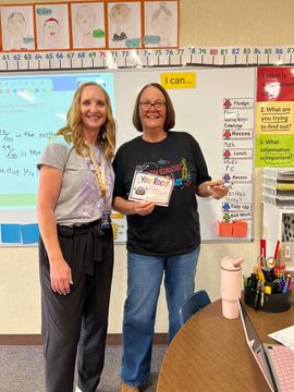 Two people smiling in a classroom, holding a certificate and standing beside a whiteboard.
