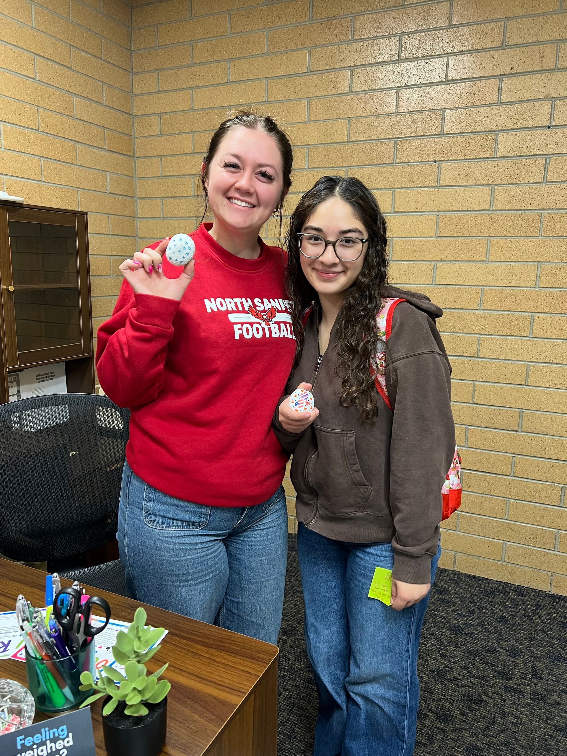 Two women smiling indoors, holding small cups, standing beside a desk and brick wall.
