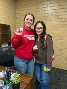 Two people standing indoors, smiling and holding small items, beside a desk and brick wall