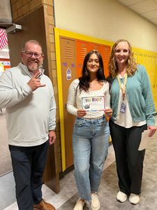 Three people smiling indoors, one holding a certificate, standing by a yellow bulletin board.