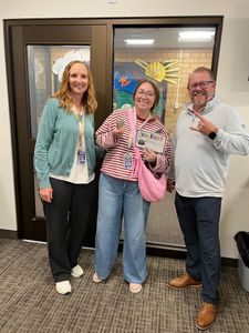 Three people smiling in front of a door, one holding a pink purse and one making a peace sign.