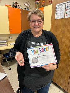 Woman in a classroom holding a colorful “YOU ROCK!” certificate and smiling.