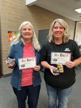 Two women holding “You Rock!” certificates and a small award, standing in a hallway.