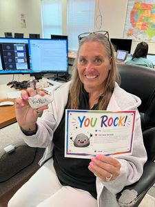 Woman holding a “You rock!” card and a small gift at a desk in an office.