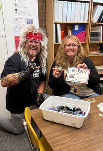 Two people at a desk holding a decorated card beside a bin of small wrapped items in a classroom.