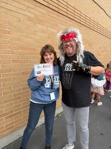 Two people smiling by a brick wall, holding a sign and making playful hand gestures.