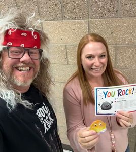 Two smiling coworkers hold “You ROCK!” cards and a small yellow treat against a stone wall.