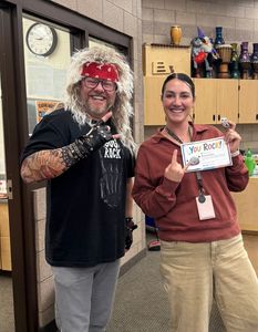 Two people smiling in a classroom, one holding a decorated card and the other wearing a red wig and costume.