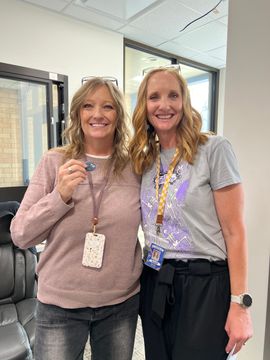 Two smiling coworkers stand in an office, wearing lanyards and casual clothes.
