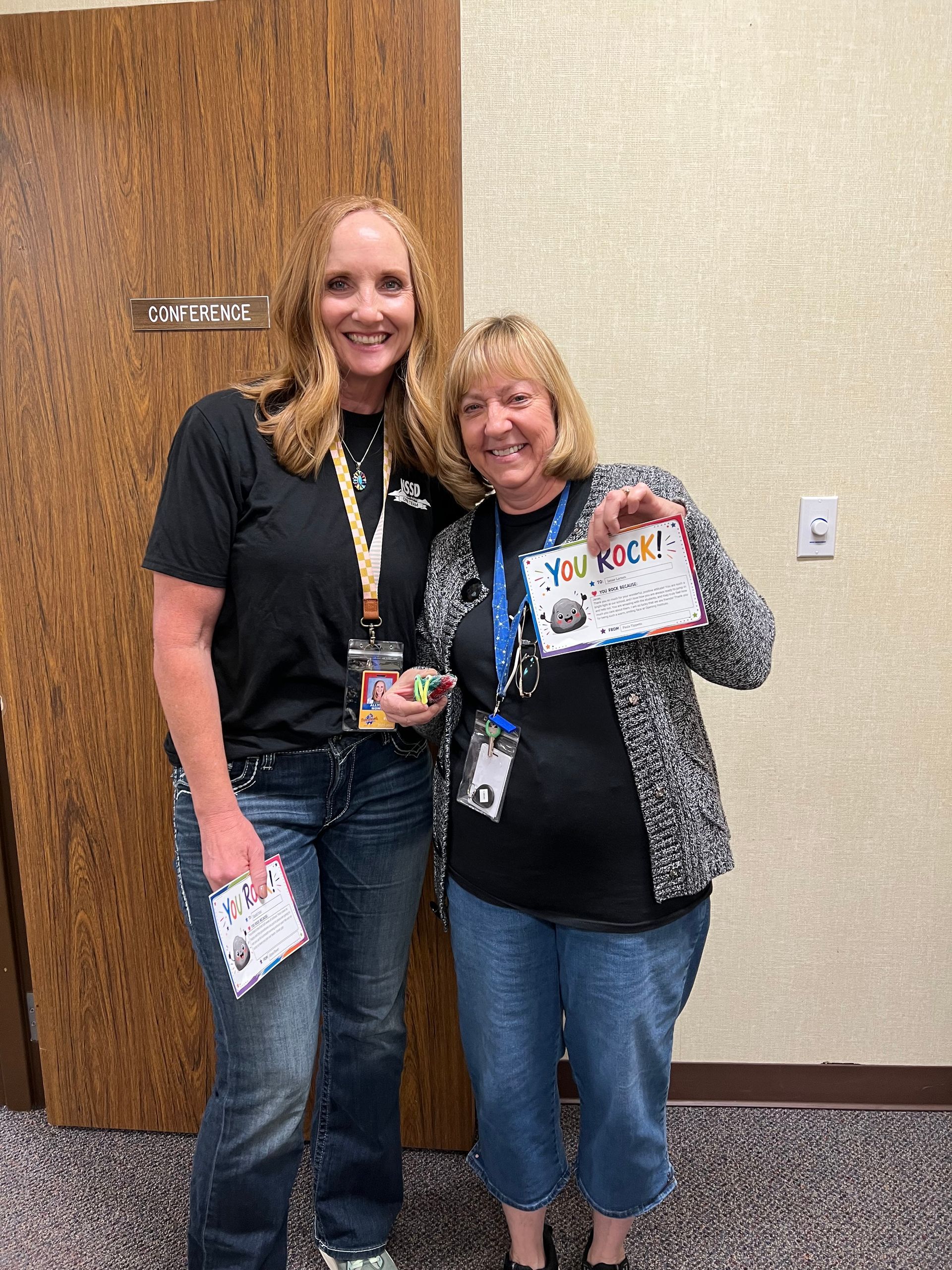 Two smiling women posing indoors, holding event materials, with lanyards against a beige wall.
