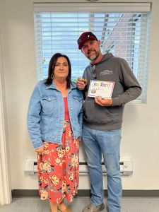 Two people stand indoors by a window, smiling; one holds a certificate or award plaque.