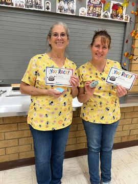 Two people in matching yellow shirts hold colorful signs in front of a school hallway display.