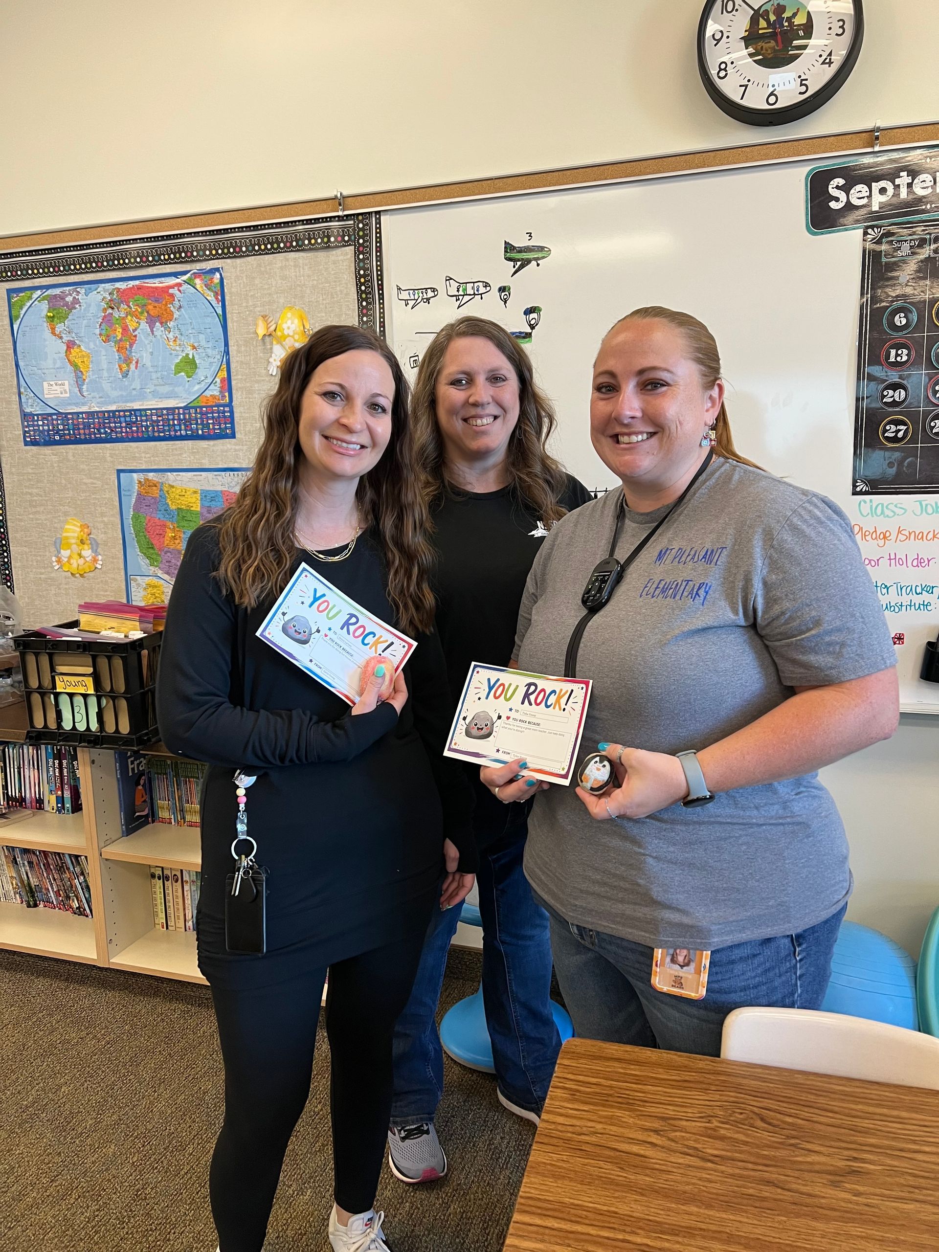 Three people smiling and holding wrapped gifts in a classroom with maps and a clock on the wall