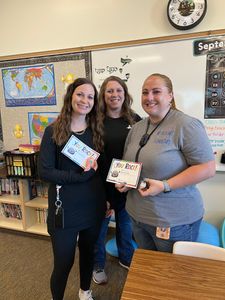 Three smiling people holding gift boxes in a classroom with a world map and wall clock.