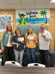 Group of four smiling people holding papers in front of a “Welcome Back Educators!” wall display.