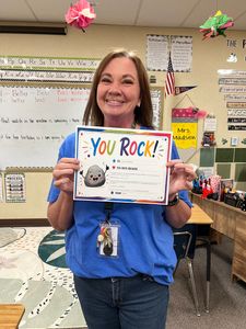 Smiling teacher holding a colorful “YOU ROCK!” award in a classroom.