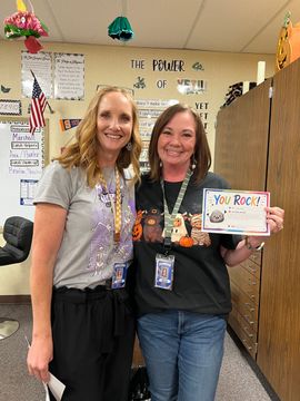 Two women posing in a classroom, smiling beside a colorful “Bye Boys” sign and decorated wall.