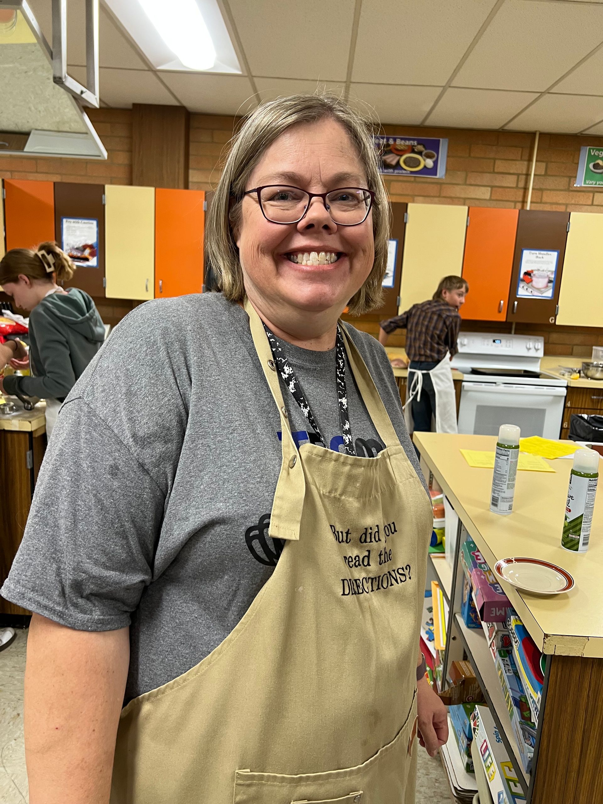 Smiling woman in a gray shirt and tan apron stands in a classroom or workshop.
