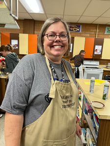 Smiling woman in gray shirt and tan apron standing in a classroom or workshop with craft supplies around her