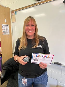 Smiling woman in a classroom holding a colorful “You Rock!” card and a small silver object