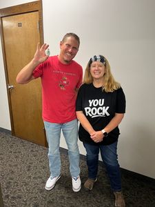 Two smiling people stand indoors, one waving, beside a door and beige wall.