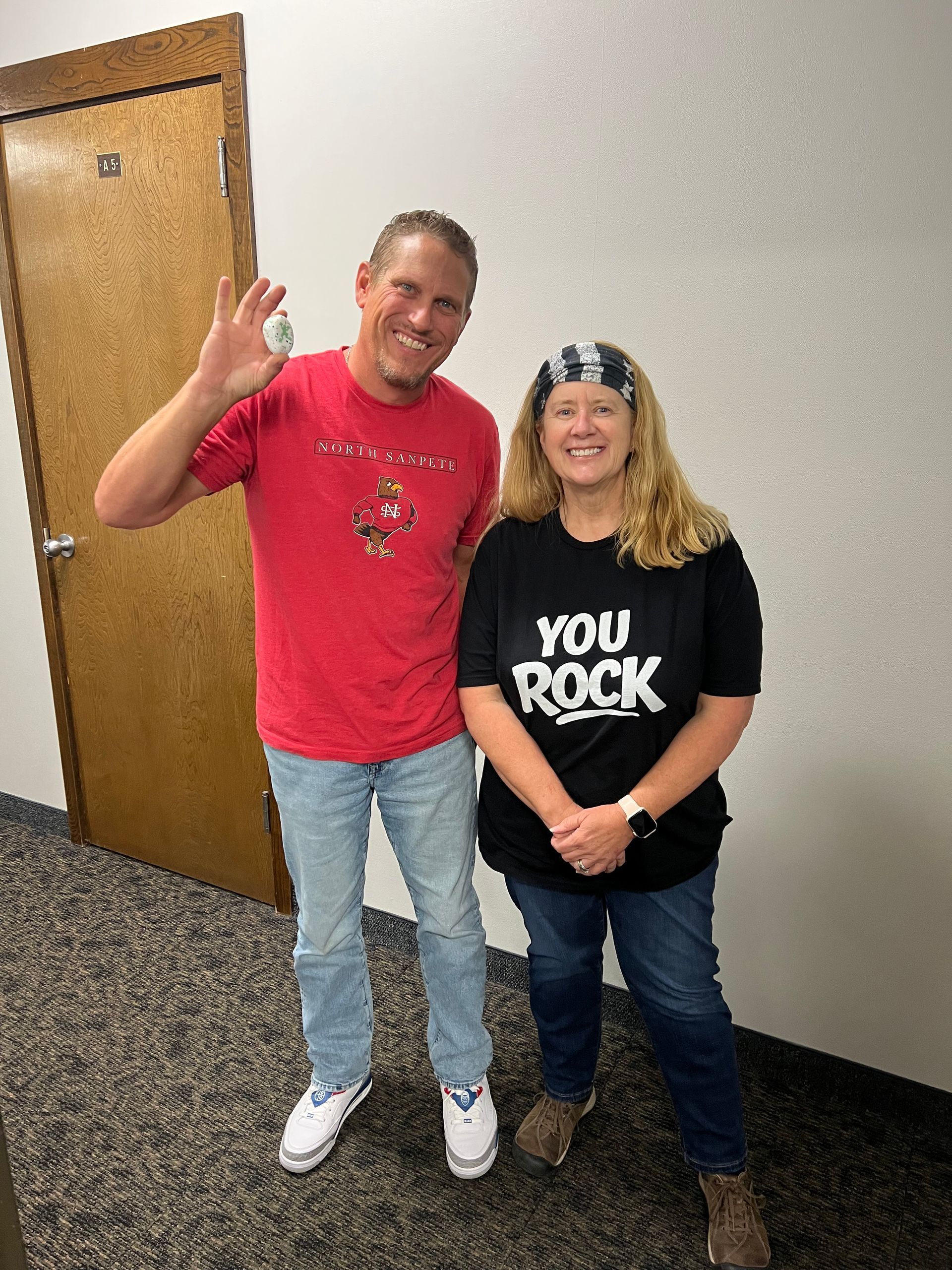 Two people standing indoors; one waves, the other wears a black “YOU ROCK” shirt.