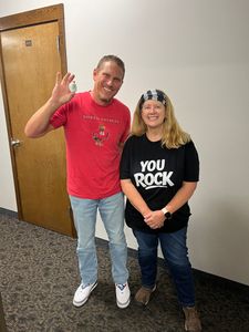 Two people posing indoors; one waves in a red shirt, the other smiles in a black “YOU ROCK” shirt.