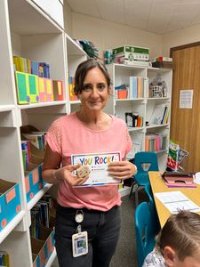 Woman in pink shirt holding a “You Rock!” card in a classroom or office with shelves of books and supplies.