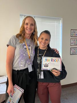 Two women smiling indoors, holding a “You Rock!” sign and a folder in front of blinds.