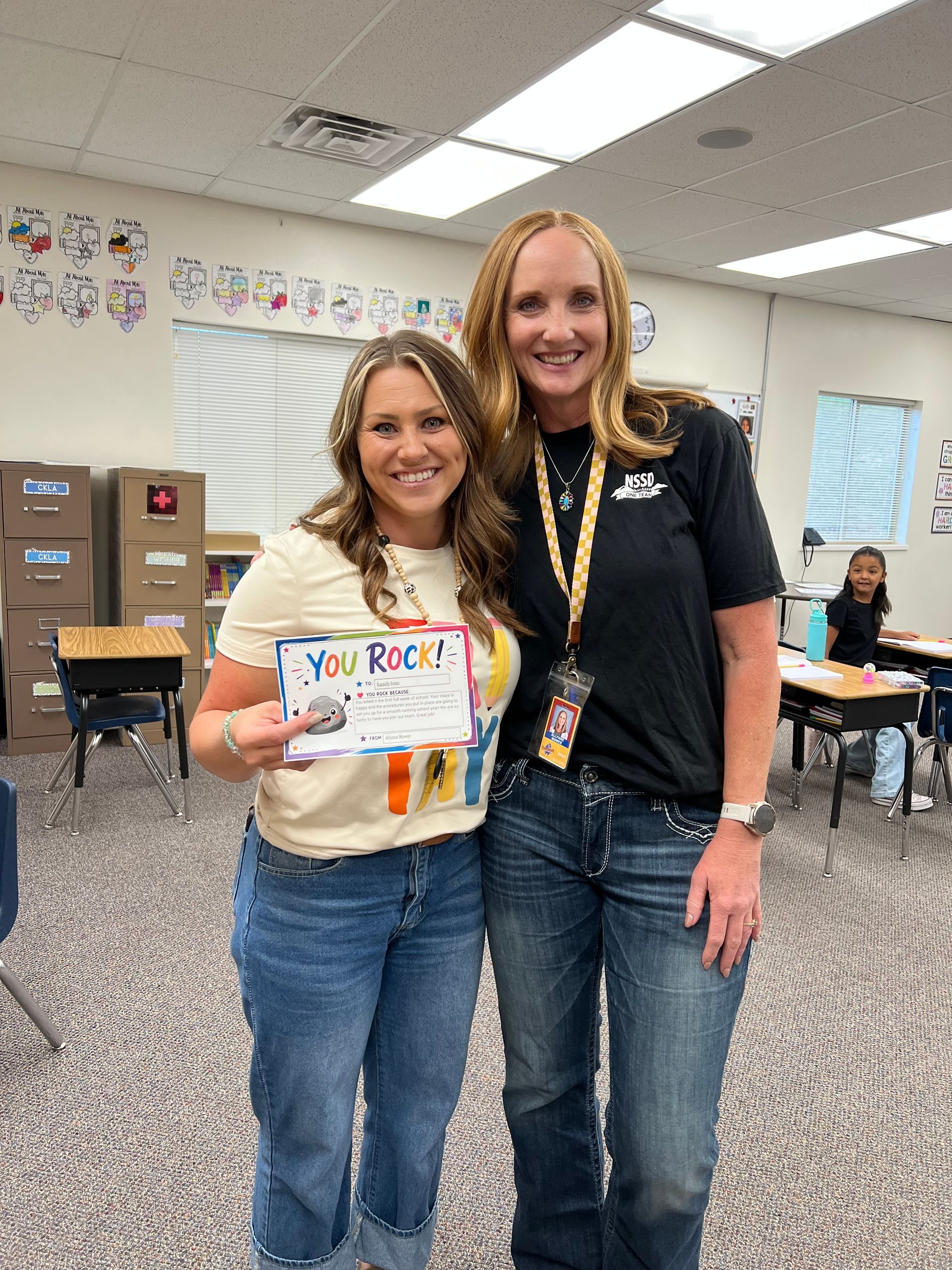 Two women smiling in a classroom, one holding a colorful poster board.