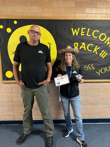 Two people stand before a black-and-yellow “Welcome Back” mural, one holding a small certificate.