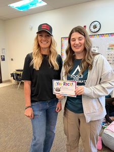 Two women smiling indoors, holding a colorful “You Rock” card in a classroom.