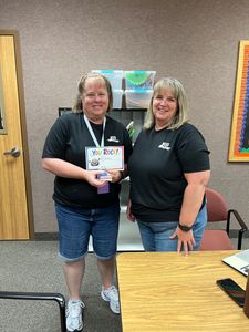 Two women in black shirts stand in an office, smiling and holding a small box.