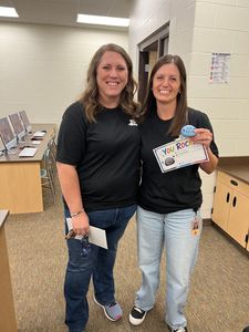 Two people smiling in an office hallway, one holding a small sign and a sheet of paper.