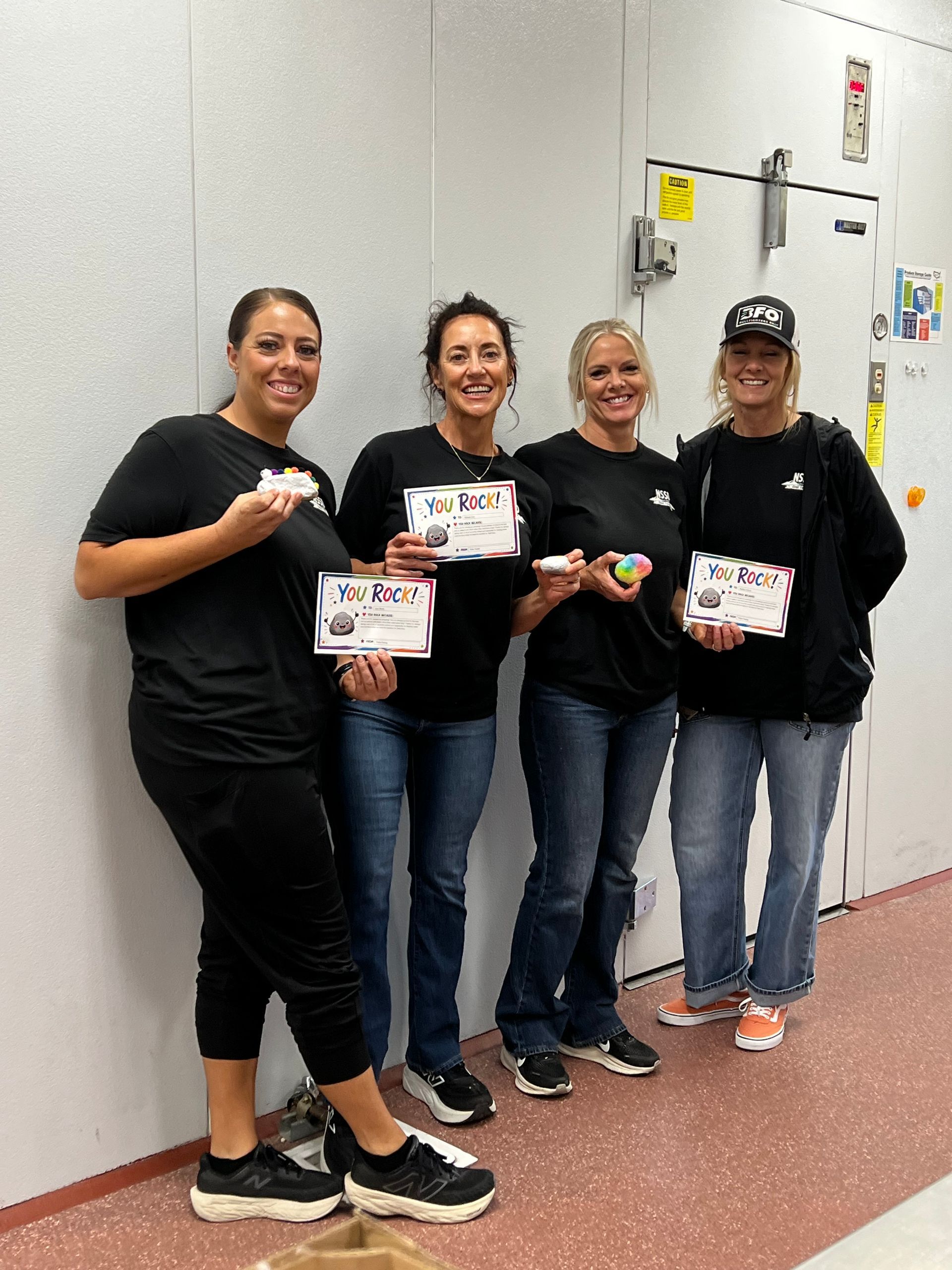 Four people in black shirts stand indoors holding small boxes and smiling near a wall panel.
