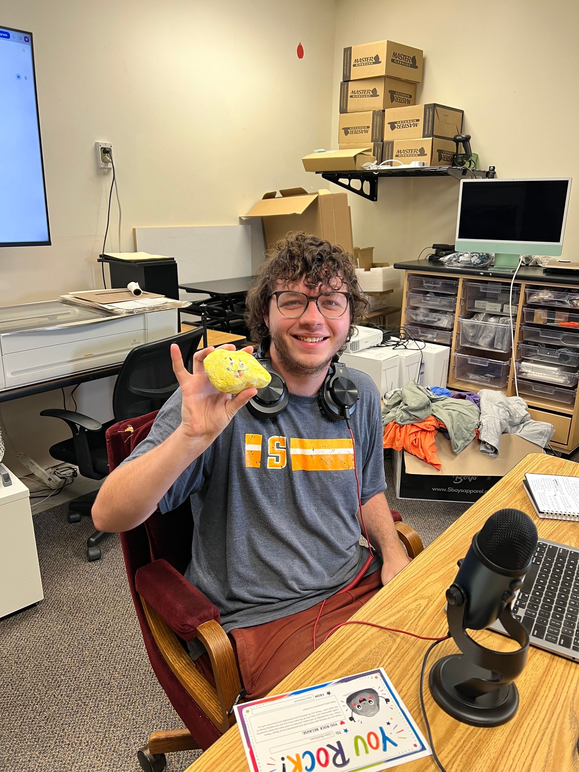 Person smiling in an office, holding up a yellow object at a desk with a microphone and computer nearby.