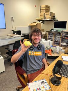Person smiling in an office, holding up a yellow object at a desk with a microphone.