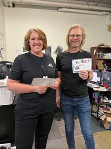 Two people standing indoors, smiling and holding a certificate or award.