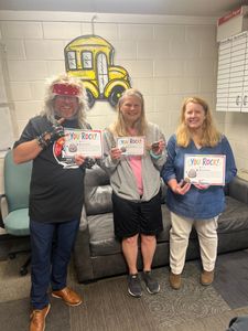 Three people holding certificates stand smiling in an office with a yellow wall mural.