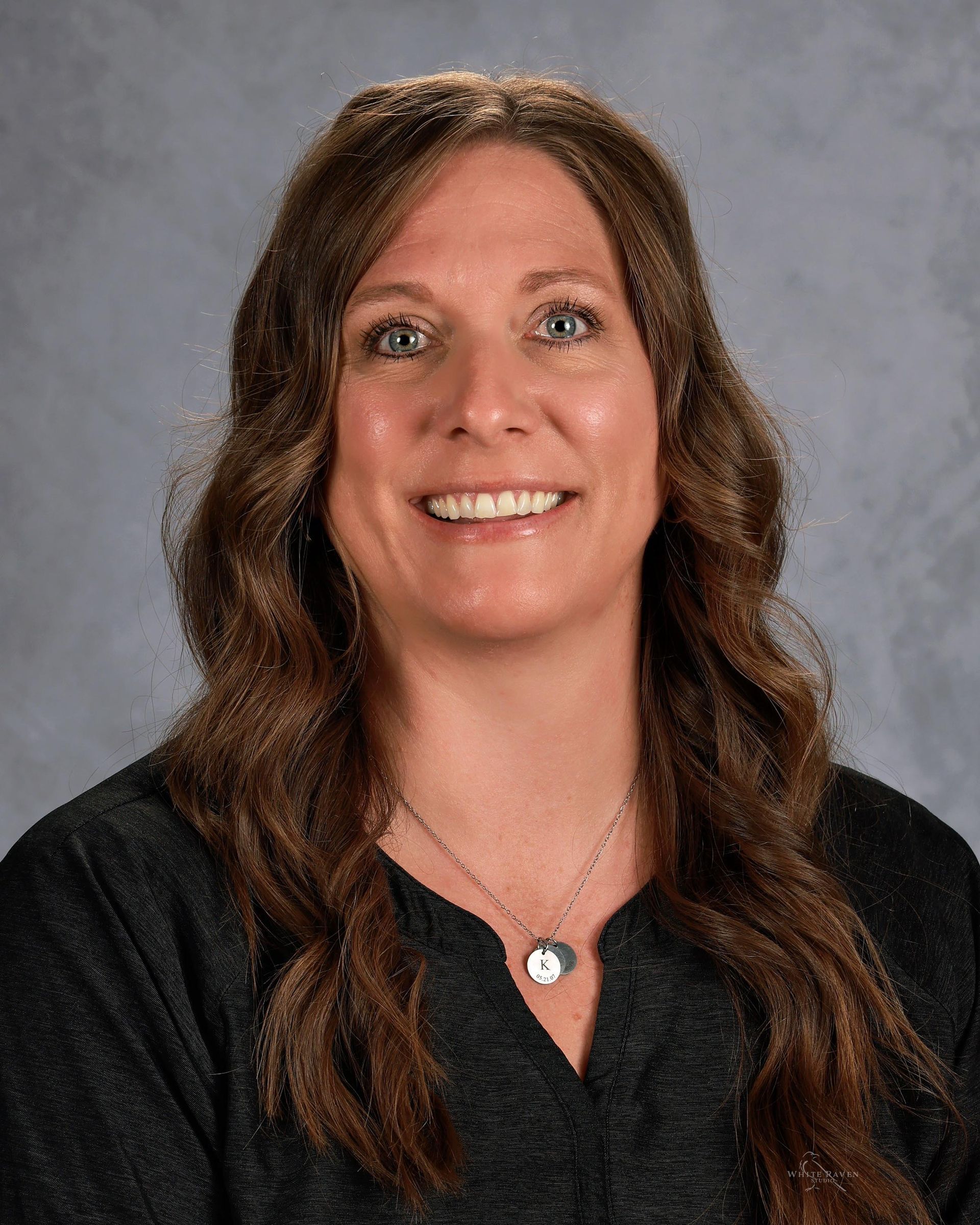 Woman with long brown hair, smiling. She wears a dark top and a necklace. Gray background.