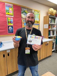 Man holding a small craft and a “YOU ROCK” sign in a classroom, smiling beside a bulletin board.