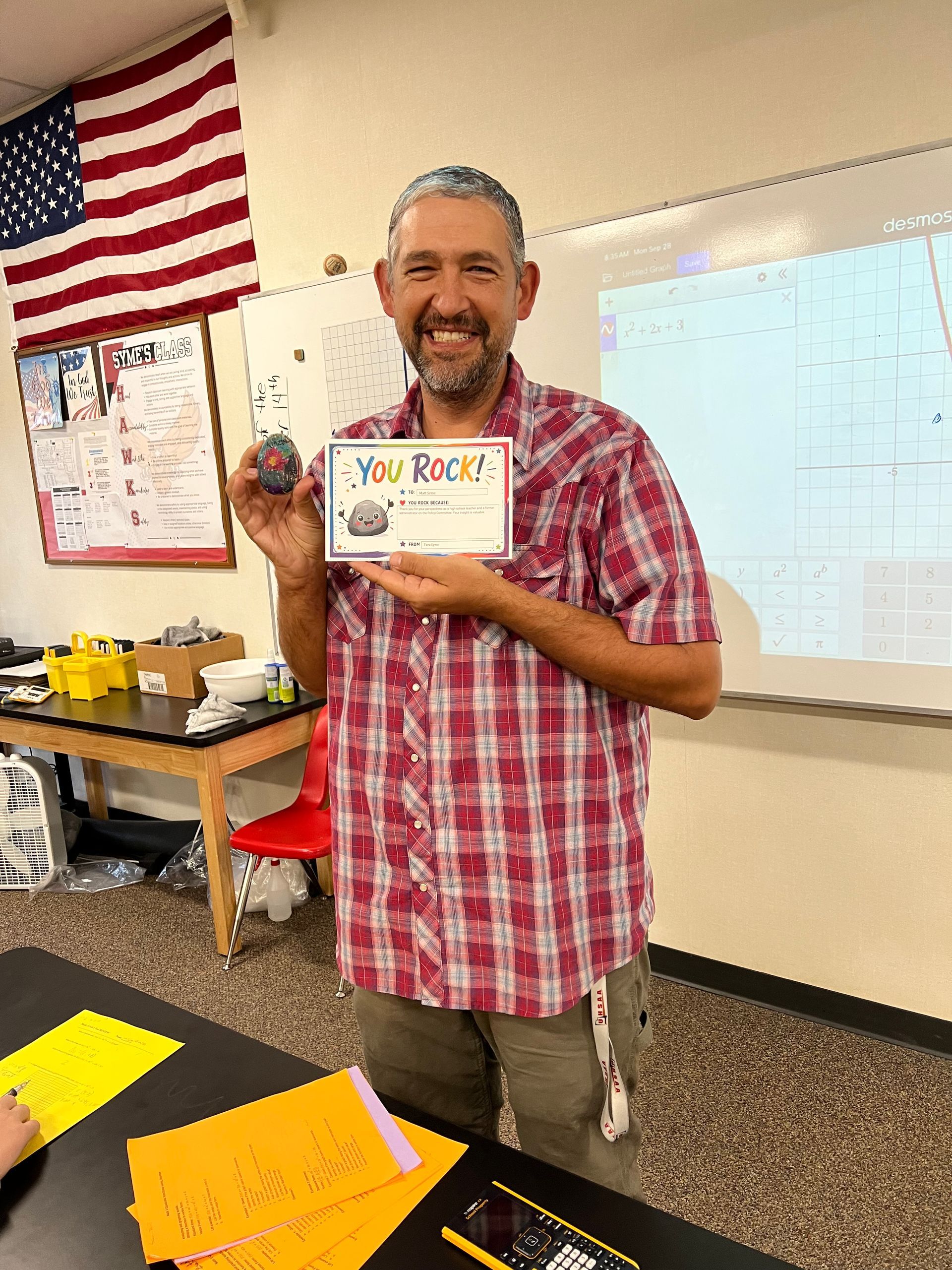 Teacher smiling in classroom, holding a “You Rock!” card beside an American flag and whiteboard.