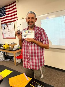 Man in a red plaid shirt holding a “You did it!” card in a classroom with an American flag.