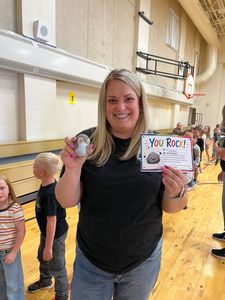 Woman in a hallway holding a colorful “You Rock!” card and a small gift, smiling at the camera.