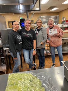 Four people standing in a kitchen, smiling behind a counter with a bowl of chopped lettuce in the foreground