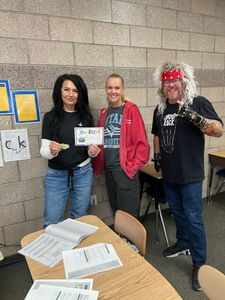 Three people holding signs stand behind a table with papers in a gym-like room.