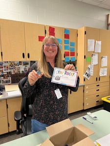Smiling teacher holding a decorated card in a classroom, with colorful cabinets and desks behind her.